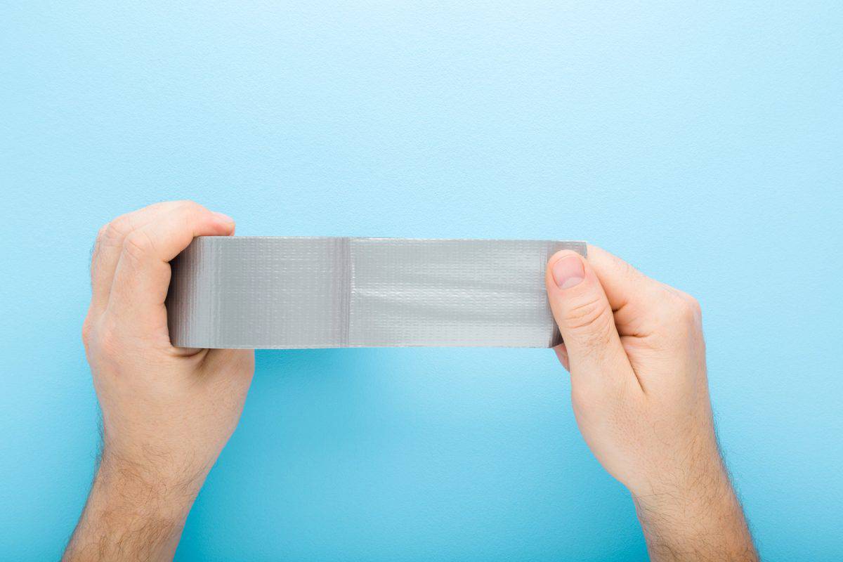 Young adult man hands stretching gray adhesive tape on light blue table background. 