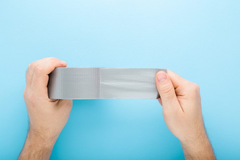 Young adult man hands stretching gray adhesive tape on light blue table background - 5 Types Of Packing Tape