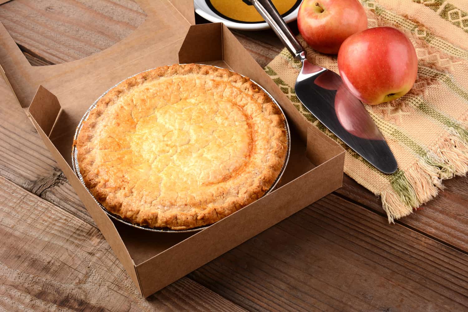 Closeup of a fresh apple pie in a bakery box on a rustic wood table. A plate, serving utensil and fresh Fuji apples are in the background. Horizontal format.