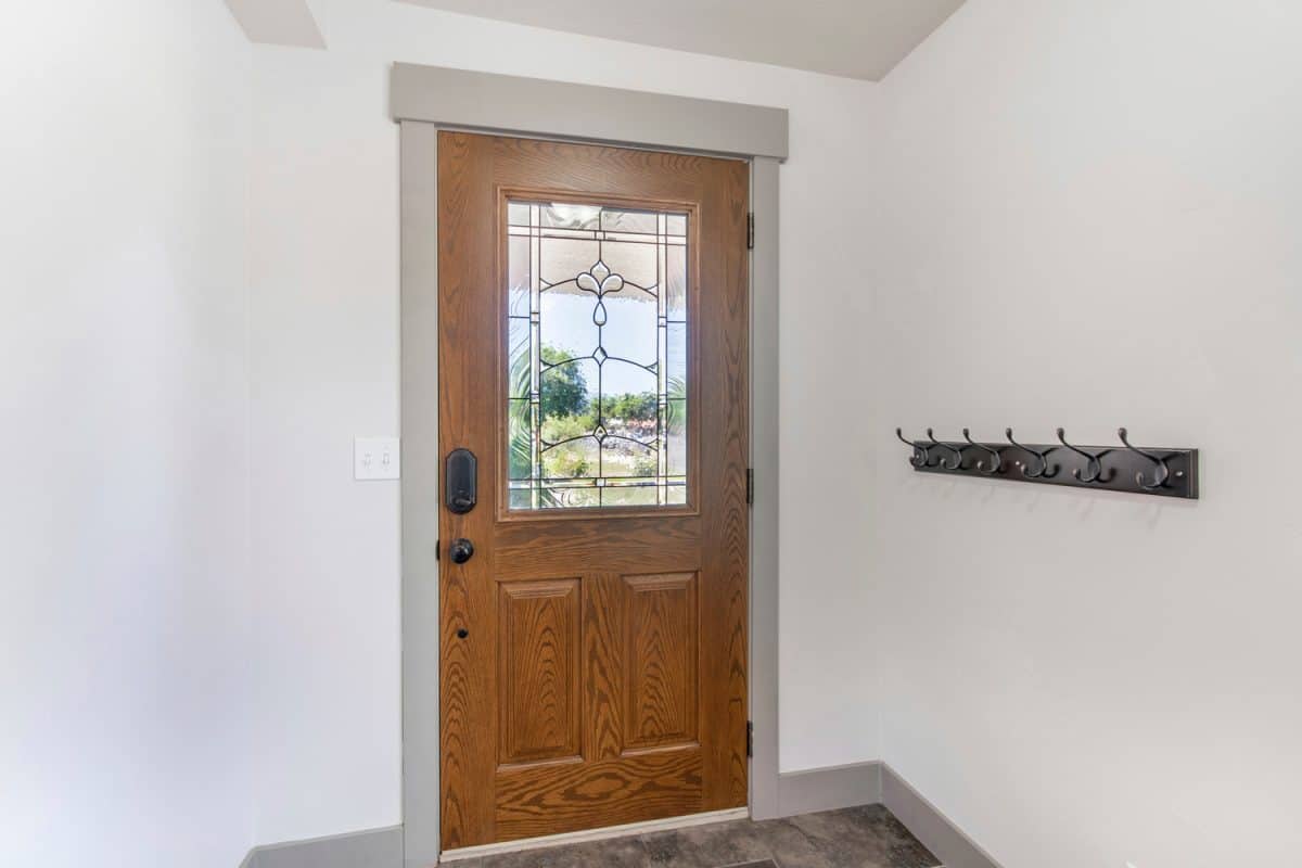 Interior of a front door with wall rail-mounted hanging hooks. There is a closed wooden door with glass panel and gray frames connected to a baseboard on a white wall.
