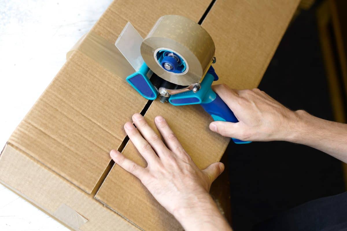 Man using a tape gun to pack a cardboard box in a factory - the mans hands and tape gun are visible

