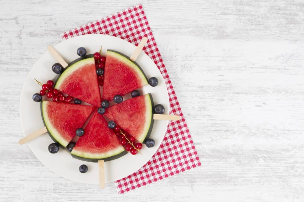 Summer concept with watermelon slice popsicles on a paper plate with blue berries and red currants on rustic wooden table.

