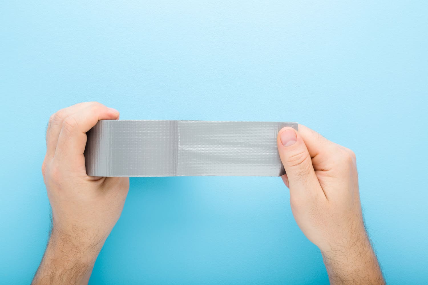 Young adult man hands stretching gray adhesive tape on light blue table background. Pastel color. Closeup. Point of view shot.